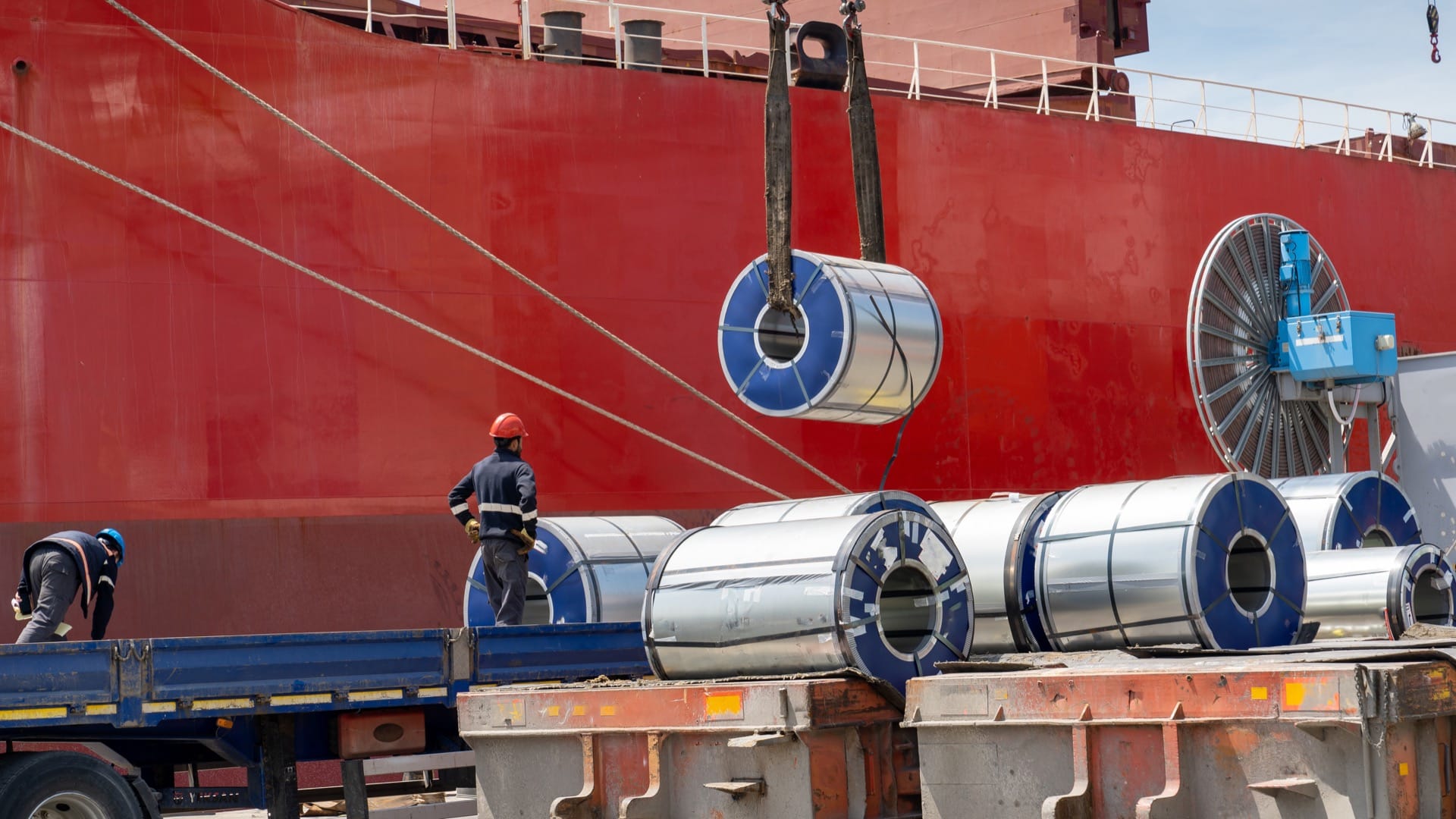Packaged coils, galvanized steel and large tin rolls are loaded onto the ship by crane from heavy duty trucks for shipment.